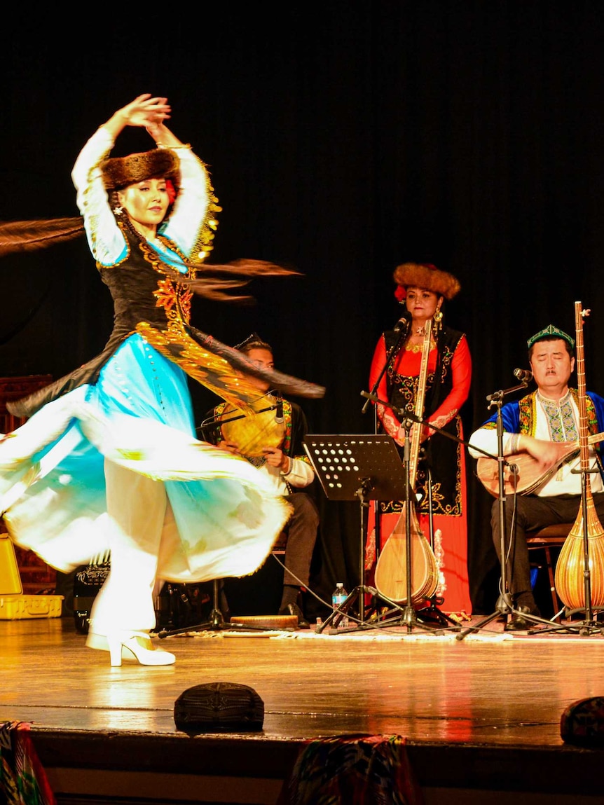 A Uyghur woman spins as she dances at a musical performance in Melbourne. Two men are playing the lute behind her on stage.