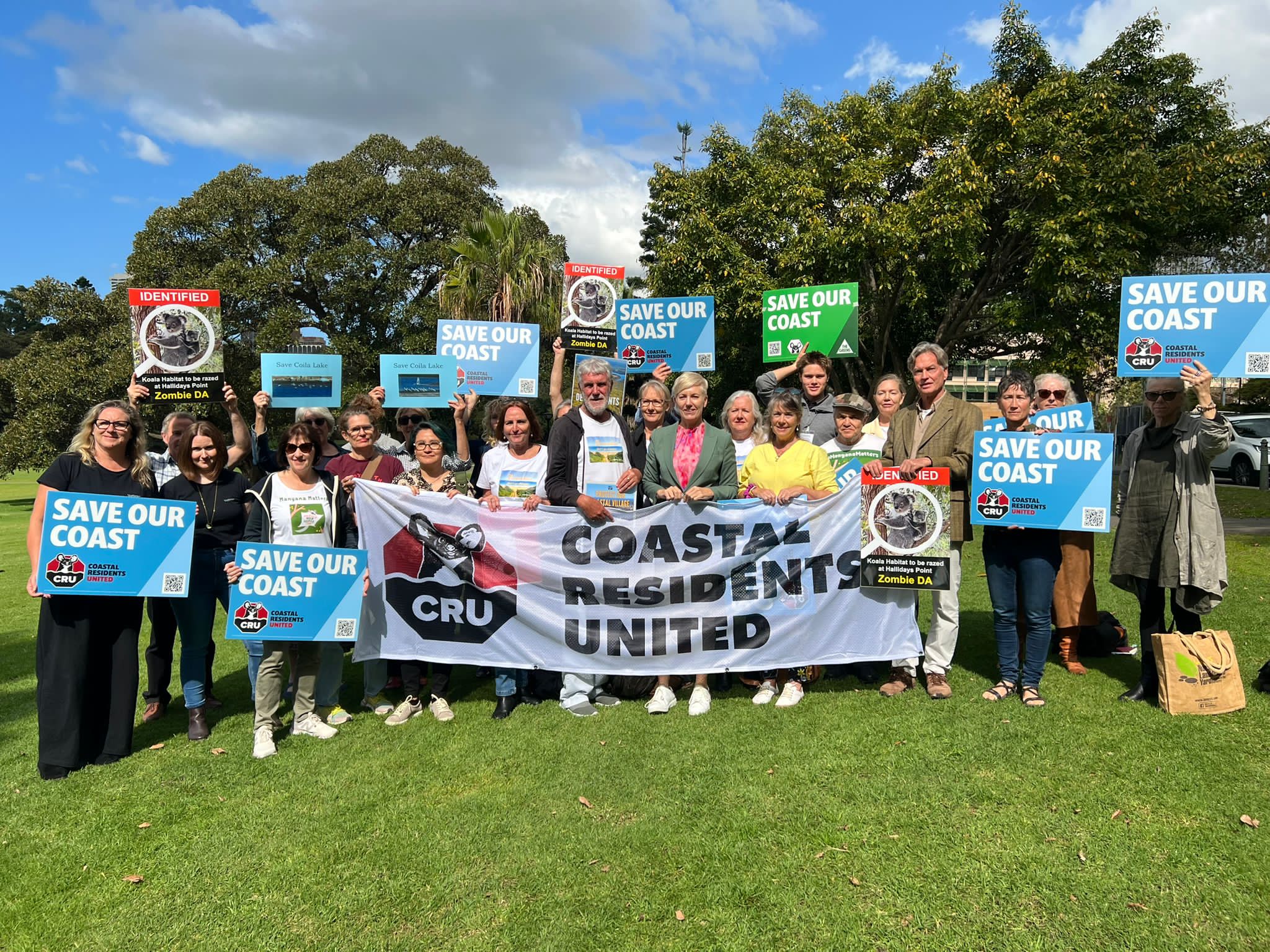 A group of people standing outdoors on a sunny day holding signs saying 'save our coast'.