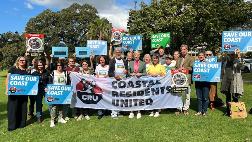 A group of people standing outdoors on a sunny day holding signs saying 'save our coast'.