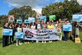 A group of people standing outdoors on a sunny day holding signs saying 'save our coast'.