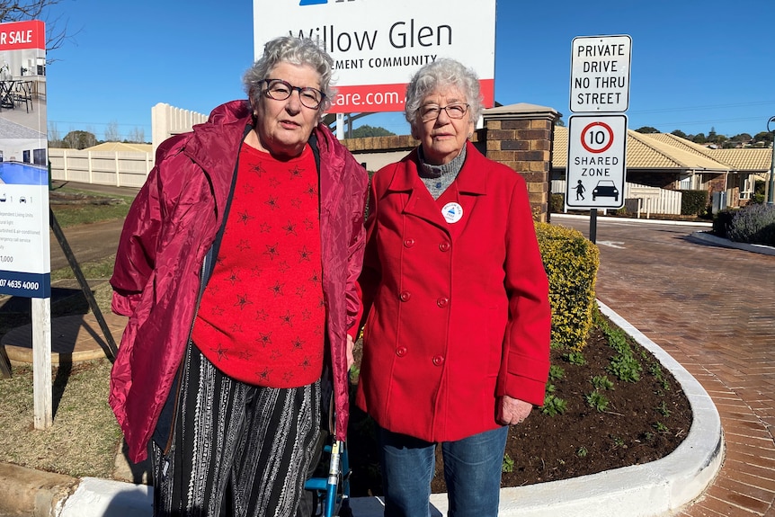 Two older women wearing red and standing in front of a sign.
