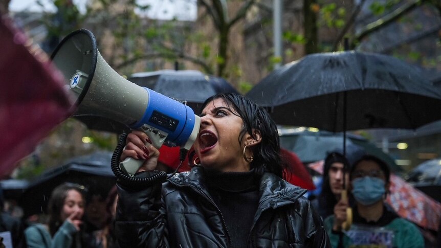 a woman speaks into a megaphone while crowds behind her hold umbrellas and signs
