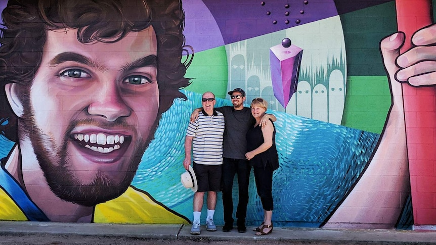 The three people stand in front of a large street-art-style mural which centres on the graphic portrait of a young smiling man.