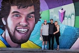 The three people stand in front of a large street-art-style mural which centres on the graphic portrait of a young smiling man.