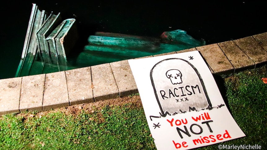 A statue is partially submerged in a pond, with a sign reading "racism, you will not be missed" next to it.