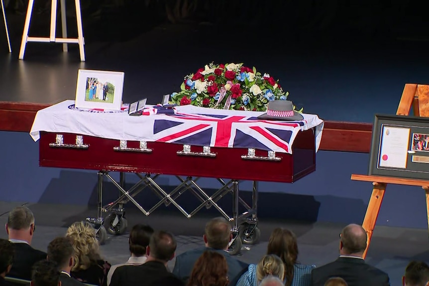 A casket draped in NSW flag with flowers, a hat, and a family photo on top of it.