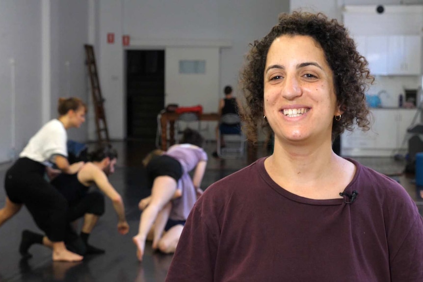 A female standing in a dance studios with dancers rehearsing in the background.
