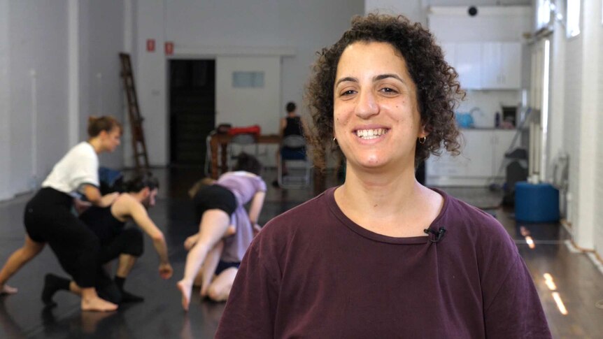 A female standing in a dance studios with dancers rehearsing in the background.