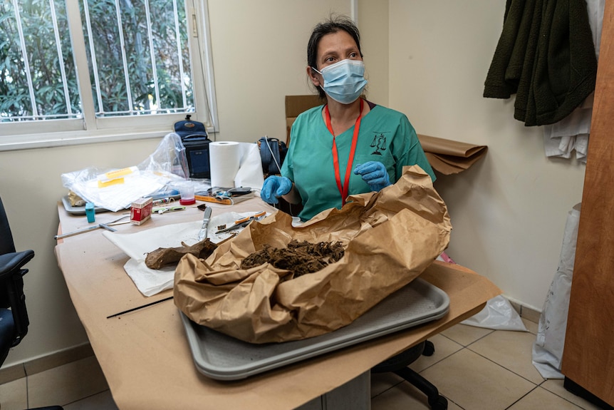 A woman wearing a mask, gloves and scrubs examines a brown paper bag holding human remains