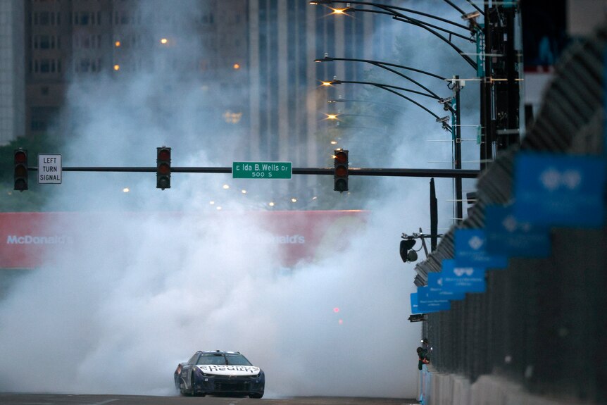 Masses of smoke rises in the air towards a street sign from a car doing a burnout driven by a NASCAR racer.