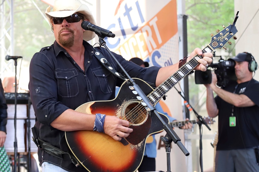 A man wearing a cowboy hat and sunglasses strums his guitar.