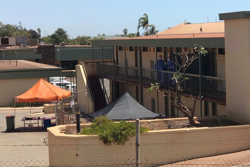 Health workers in full-length PPE on the balcony of the Hedland Hotel with an orange canopy in the car park below.