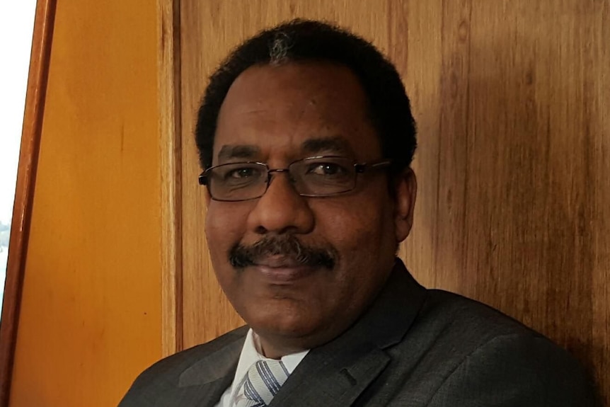 A moustachioed middle-aged African man with glasses and suit smiles and looks to camera with wooden wall behind. 