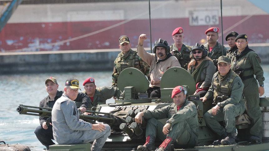 A group of twelve people in military fatigues sit atop a tank with the central figure holding his fist in the air