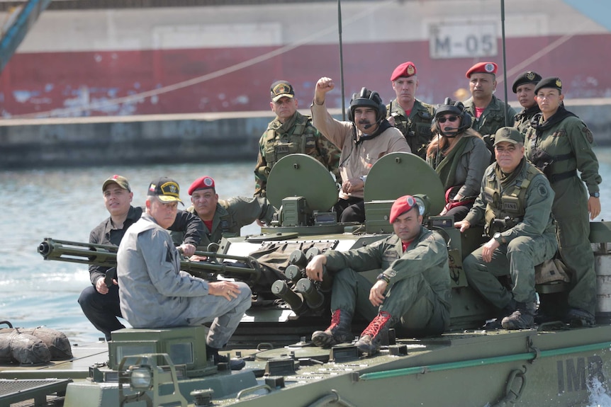 A group of twelve people in military fatigues sit atop a tank with the central figure holding his fist in the air