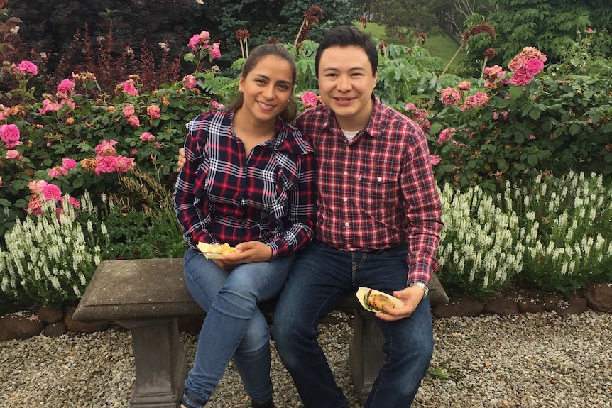 A smiling, youngish couple sit on a bench in front of a flower garden. Both wear checked flannel shirts and jeans.
