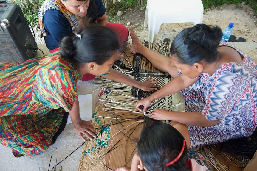 Several women working on weaving a mat while sitting on floor.