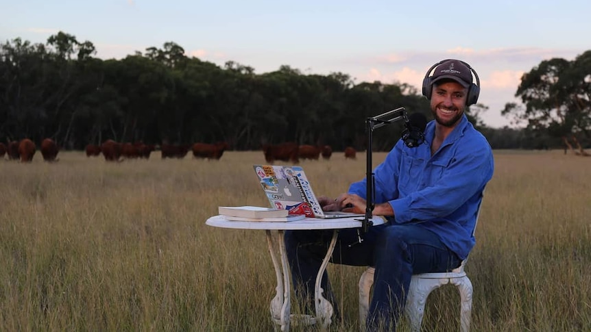 A man in his 20s sits in a field with a microphone and headphones.