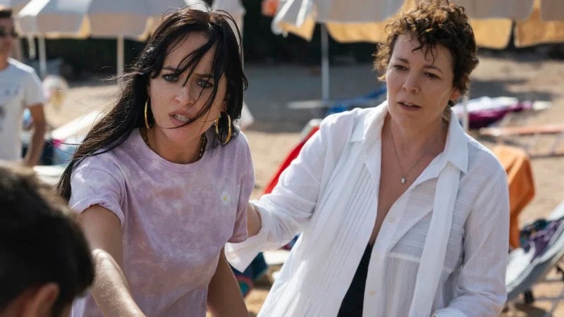 Olivia Colman in a white shirt on the beach standing next to another woman in a purple shirt with long hair and gold earrings.