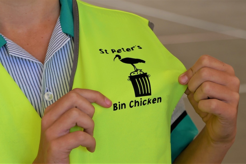 Hands holding out the corner of a high-vis yellow vest with the logo 'St Peter's Bin Chickens' around a bin and ibis drawing.
