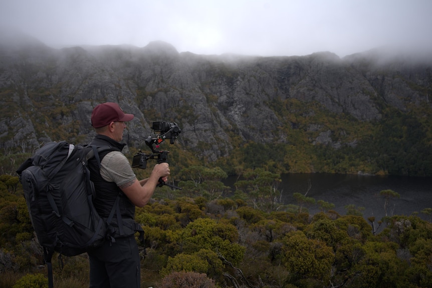 Man wearing hat and carry large backpack, filming a rugged mountain covered in fog with misty lake at the bottom. 