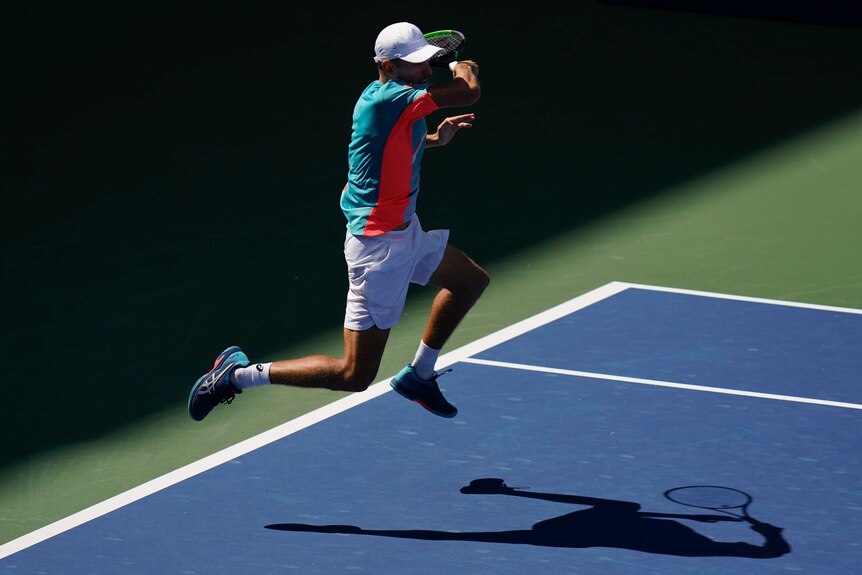A tennis player leaps off the ground to hit a ball mid-air as his shadow can be seen on the court.