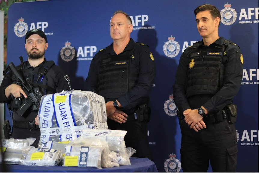 Three armed men wearing black uniforms stand behind a table full of seized drugs at a press conference.