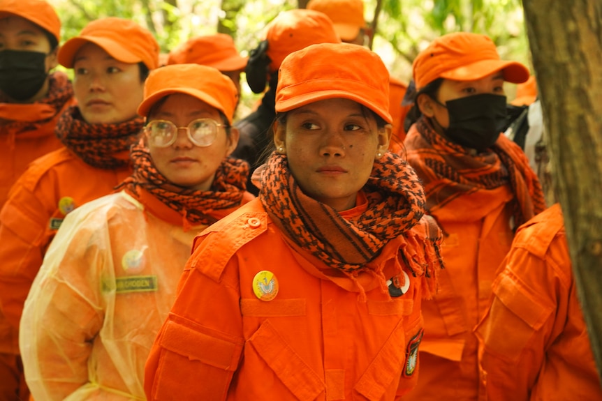 A group of women stand in a forest wearing orange uniforms and caps.