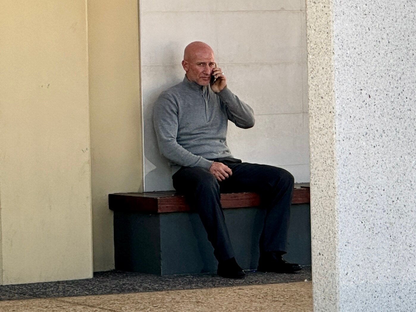 A bald man sits on a beach outside a courthouse holding his phone to his ear
