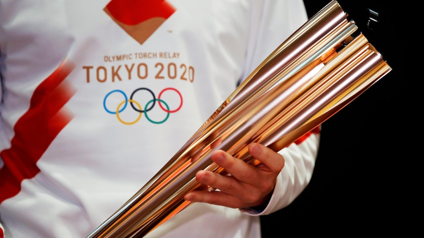 A person in a white t-shirt bearing the Tokyo Olympics logo holding the golden Olympic torch