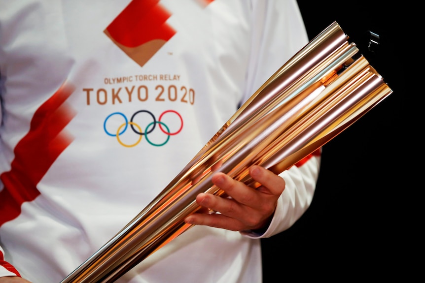 Una persona con una camiseta blanca que lleva el logo de los Juegos Olímpicos de Tokio sosteniendo la antorcha olímpica dorada