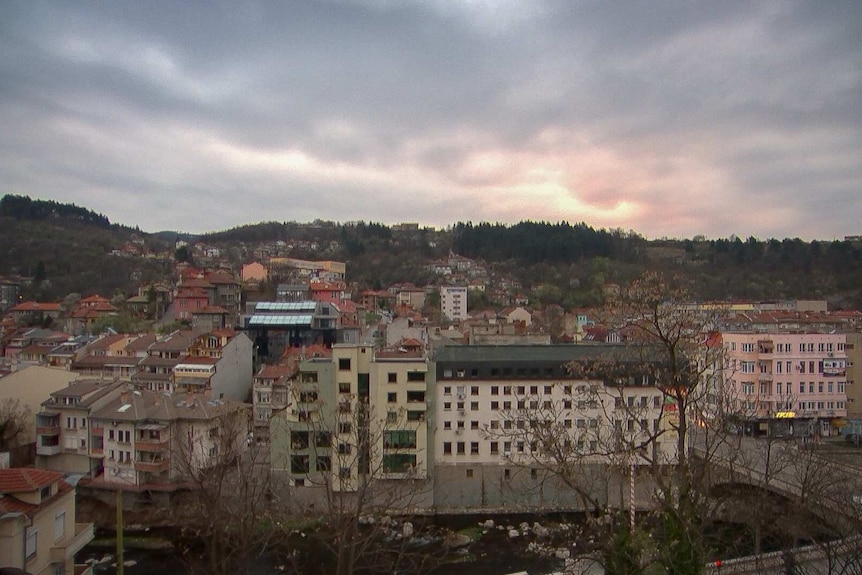 Image shows residential buildings across a river with a hill in background. The sky is great and cloudy.