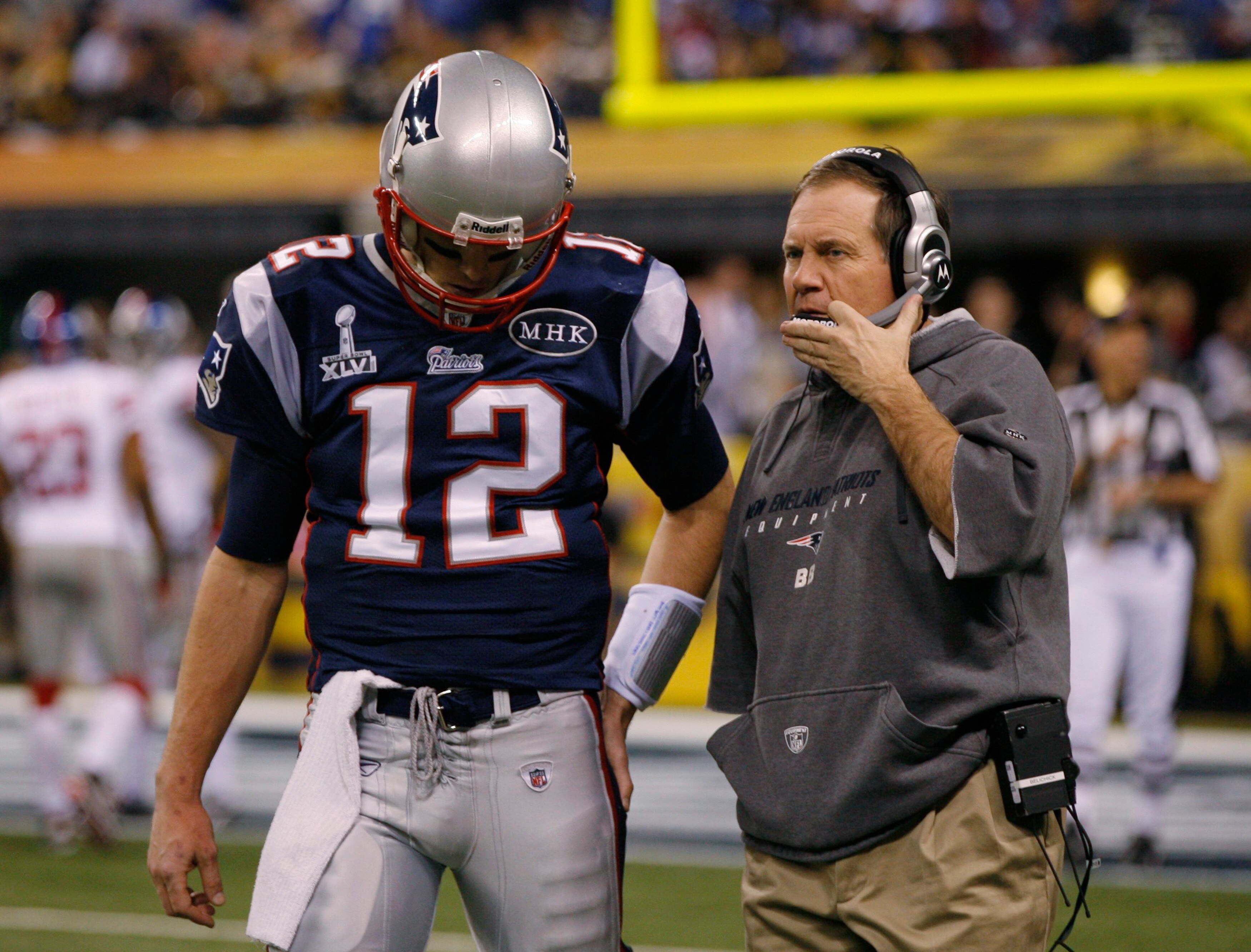 A tall NFL player in a dark blue jersey with the number 12 talks to an older man in a grey jumper who's wearing a headset.