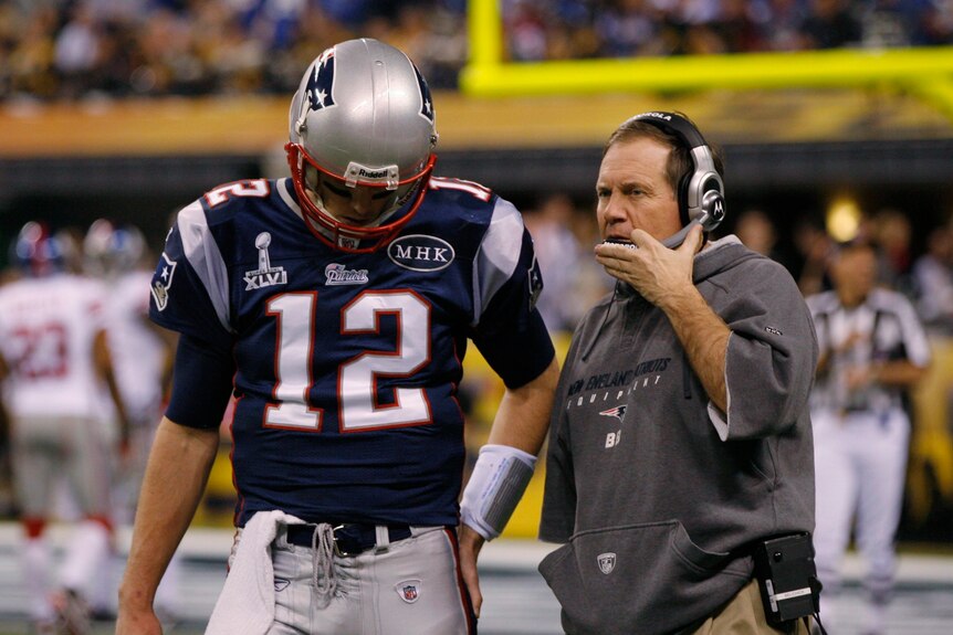 A tall NFL player in a dark blue jersey with the number 12 talks to an older man in a grey jumper who's wearing a headset.