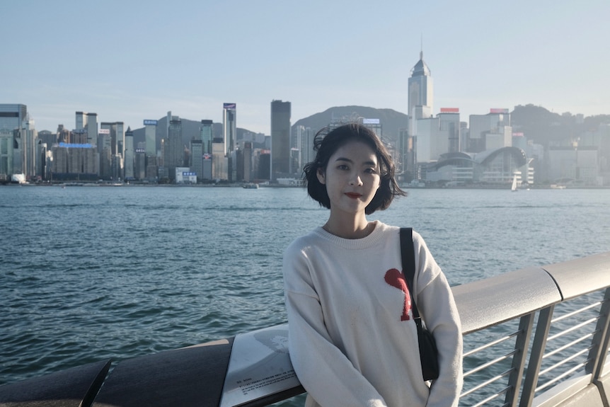 A woman in a white jumper stands in front of a harbour, looking at the camera.