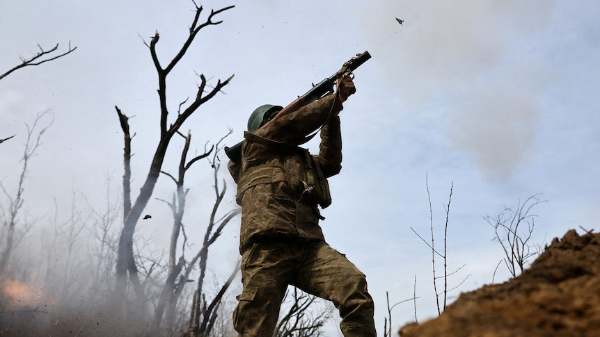 A soldier with a green helmet and camoflague fatigues fires an RPG from his shoulder while standing in a muddy trench.