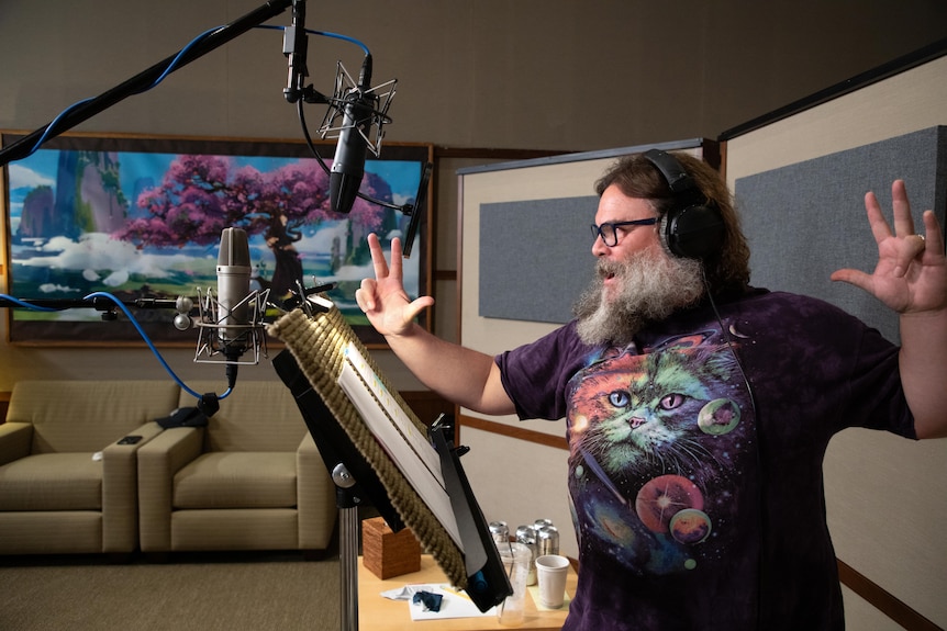 Jack Black performs kung fu gestures as he speaks into a microphone in a recording studio with animation on screen behind.