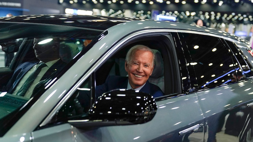 An elderly white man beams happily from the driver's seat of a shiny new car under lights.