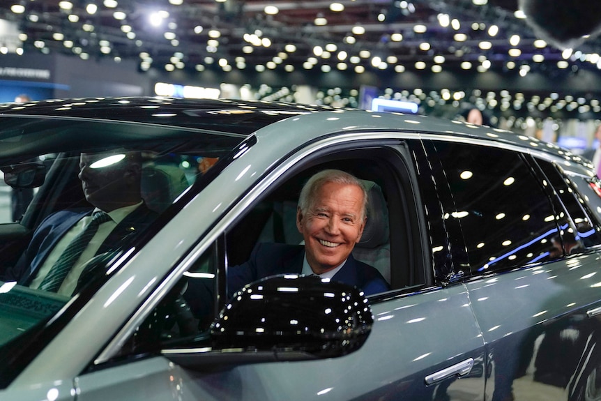 An elderly white man beams happily from the driver's seat of a shiny new car under lights.