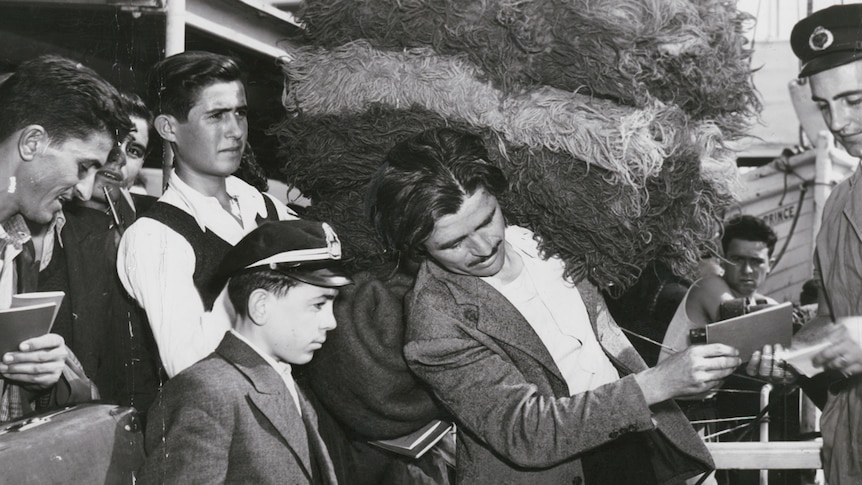 A black and white image shows a group of men in suits carrying bags and immigration documents while a man carries wool blankets.