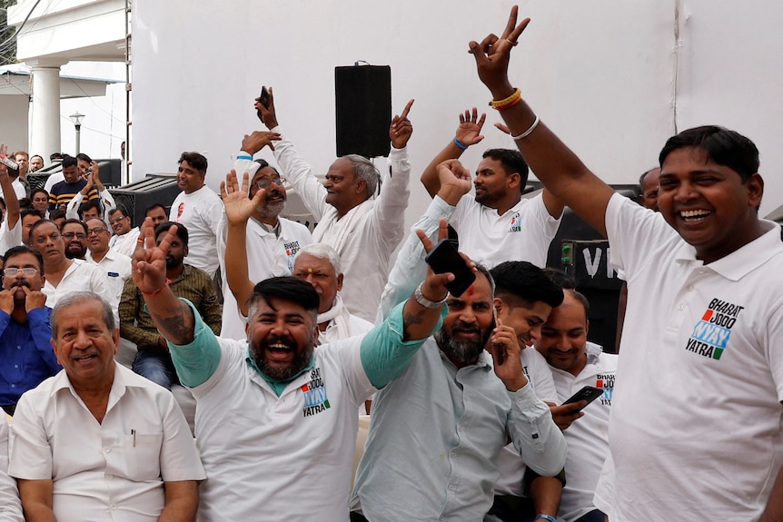 A group of men in white shirts raise their arms in the air, some making peace signs, and smiling widely