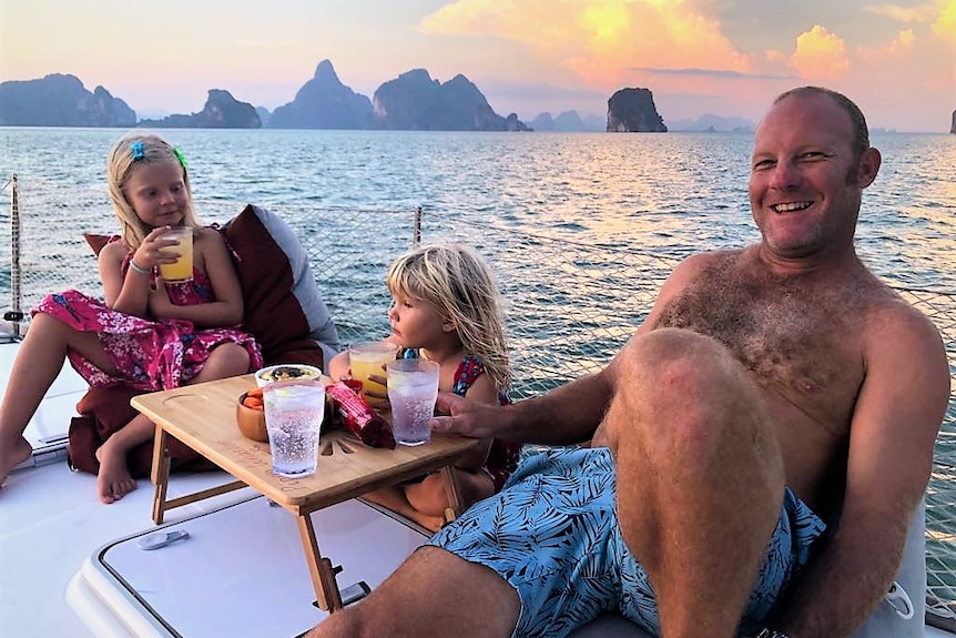 Two young girls and their dad sitting on a boat having drinks at sunset with scenic Phuket in background