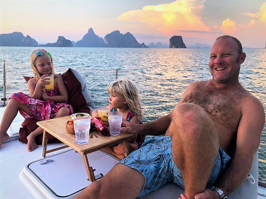 Two young girls and their dad sitting on a boat having drinks at sunset with scenic Phuket in background.