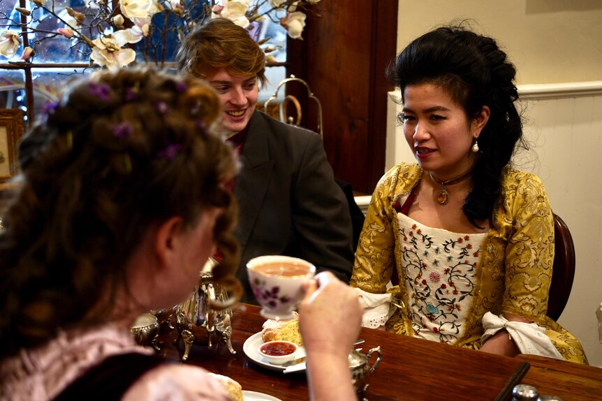 Two women and a man in old-fashioned costumes have tea indoors