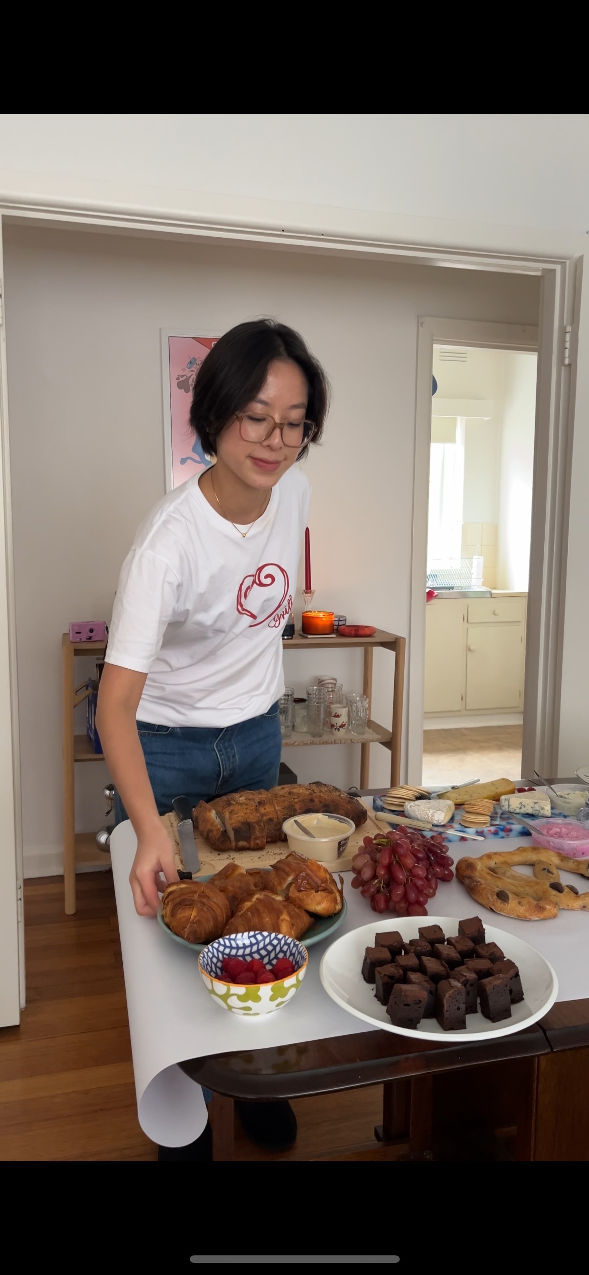 A young woman in jeans and a white printed tee standing over a table of laid out pastries, fruit and cheese. 