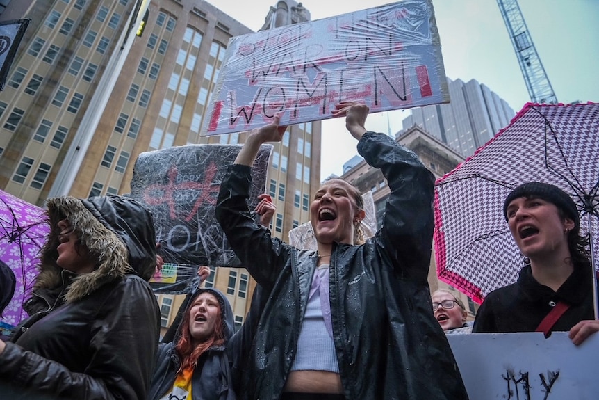 Группа протестующих держит транспаранты и зонтики на митинге протеста против выбора в Сиднее.