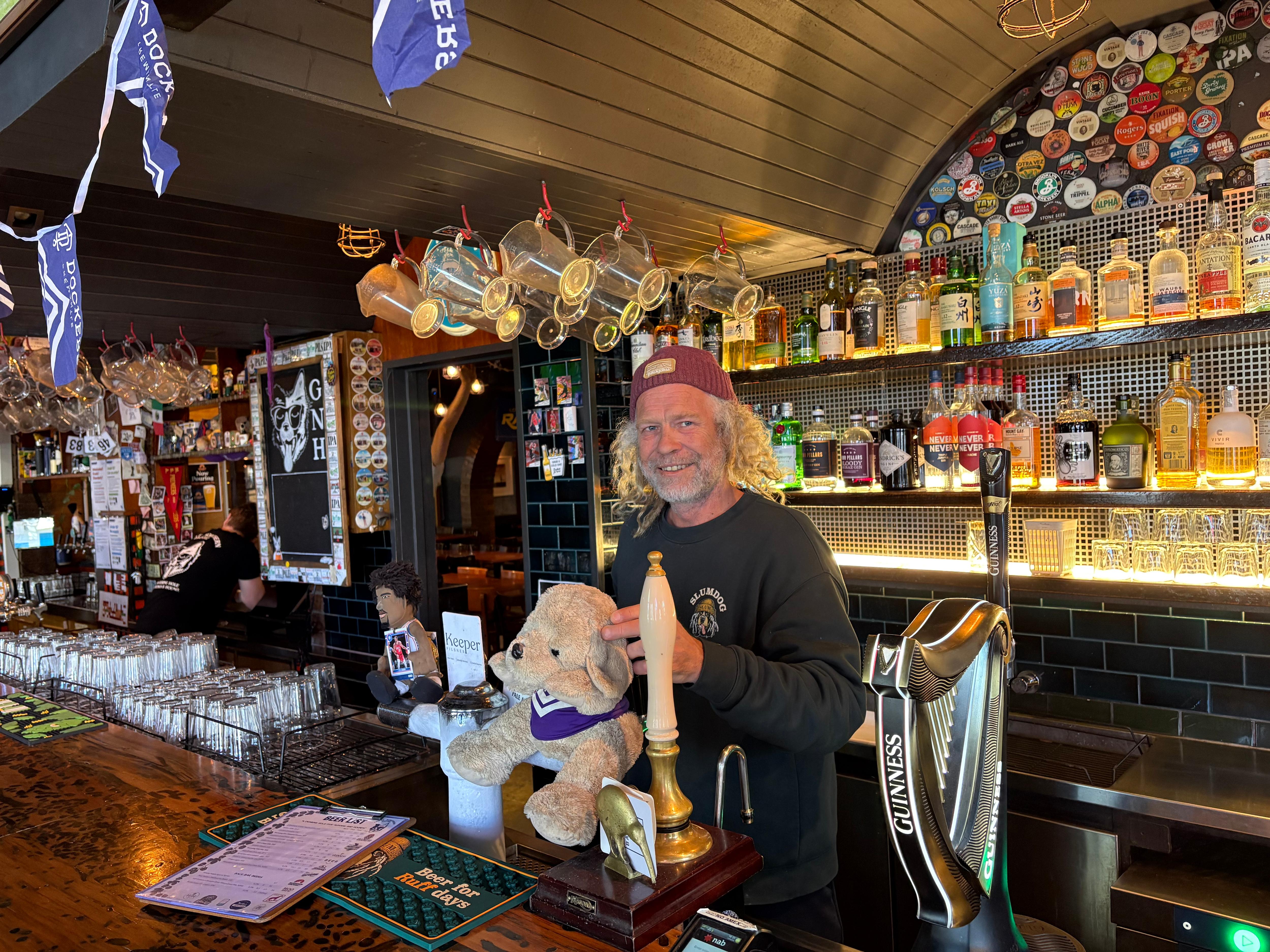 Man with long grey hair wearing a beanie standing behind a bar with Fremantle decorations around it.