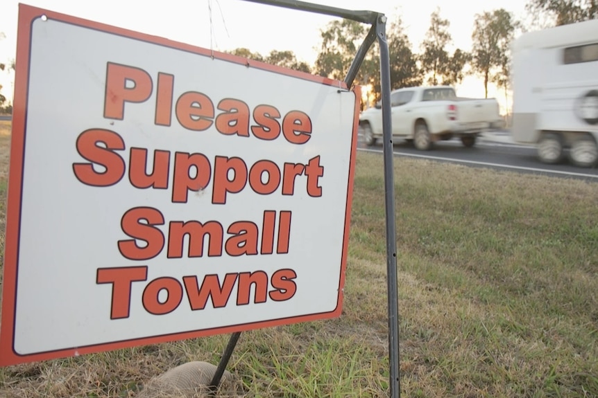 A white sign with red writing that reads 'please support small towns' sits on the side of a road