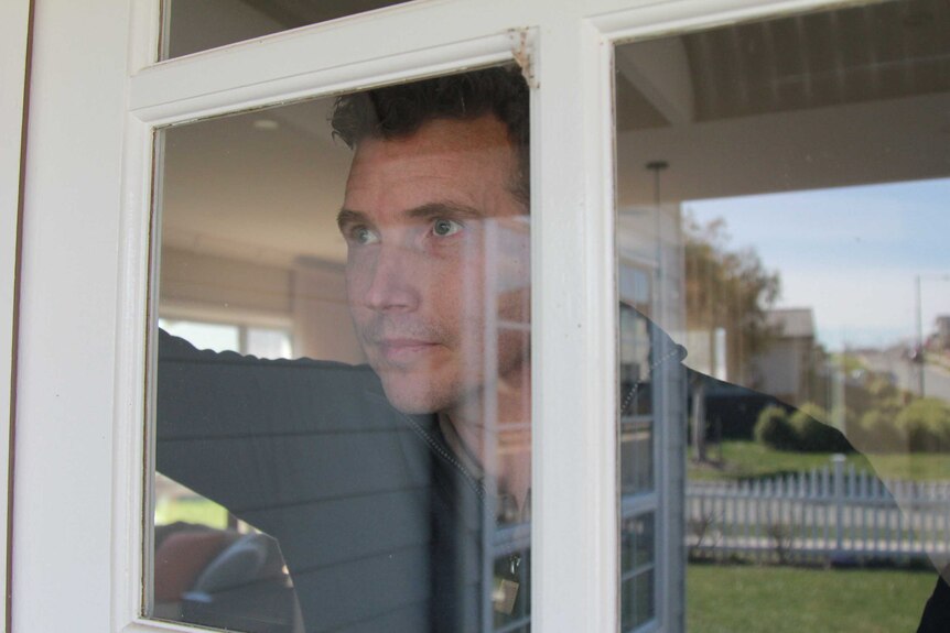 A young man looks forlornly out of his living room window.