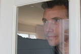 A young man looks forlornly out of his living room window.